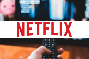 A person's hand is holding a black remote control, likely for a streaming device, with their thumb resting on the circular navigation pad. The remote has various buttons, including volume, mute, play/pause, and home.

In the background, a blurred television screen is visible, displaying colorful content, possibly a movie or show with warm tones of orange and blue.