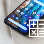 
A close-up, angled shot of a smartphone lying on a light-colored surface, possibly wood or fabric. The phone's screen is visible, displaying a grid of various app icons, suggesting a home screen. The icons include standard applications like a browser, camera, settings, and social media apps.

In the bottom right corner of the image, a partially transparent, white overlay icon of a calculator is visible.