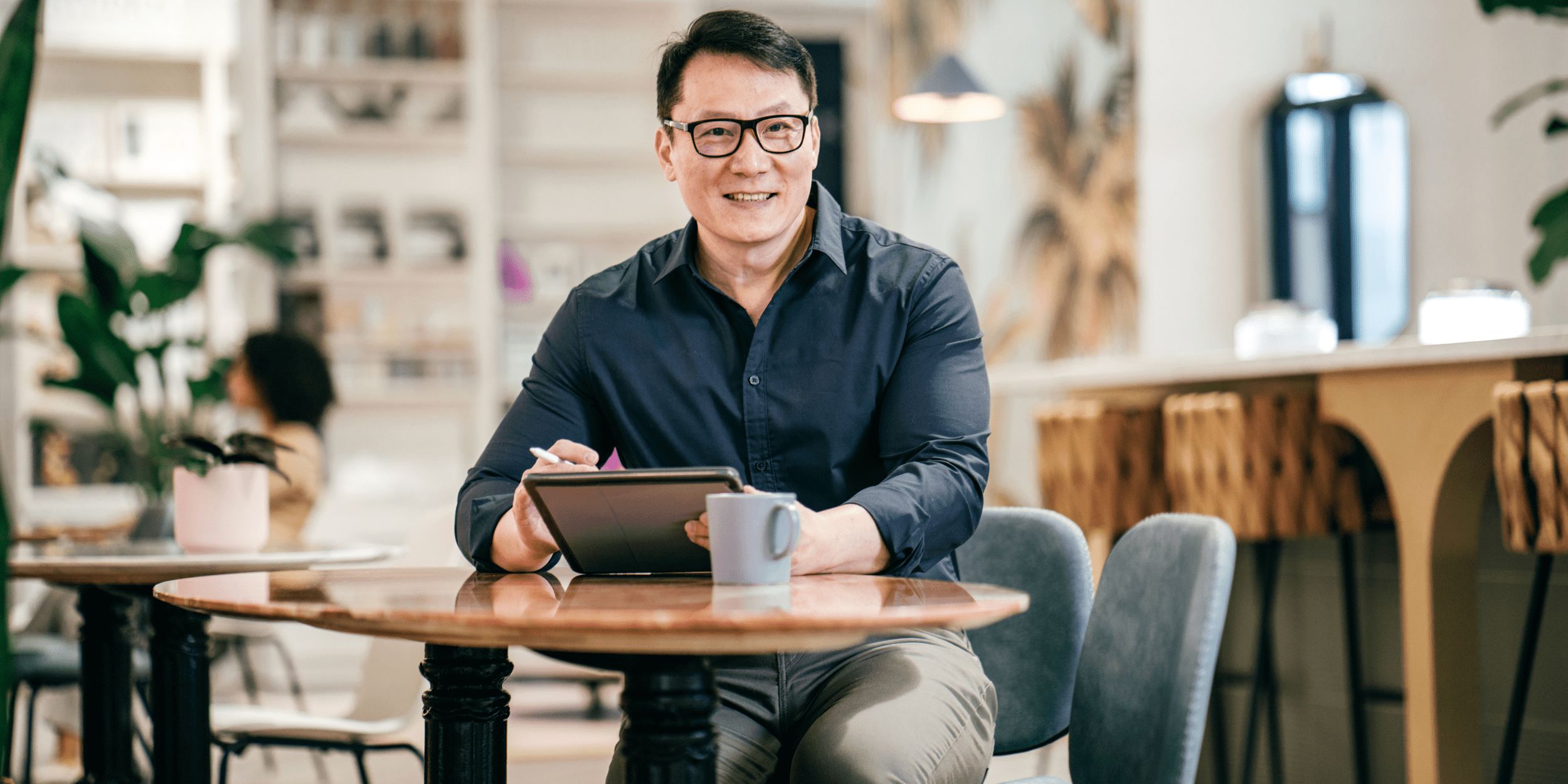 A smiling Asian man wearing glasses and a dark collared shirt is seated at a round wooden table in what appears to be a café or co-working space. He is holding a stylus in his right hand and resting his left hand on a tablet, which is also on the table. A light blue mug is next to the tablet. The background is softly blurred but shows shelves, plants, and other tables and chairs, with another person partially visible in the distance on the left. He is looking directly at the camera.